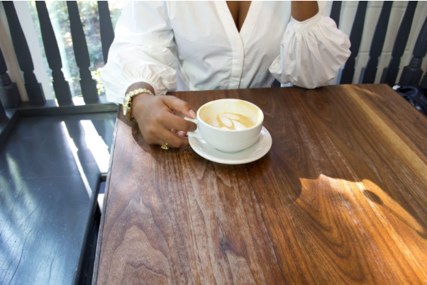 pessoa negra sentada em uma mesa de madeira segurando uma xícara de café com espuma.