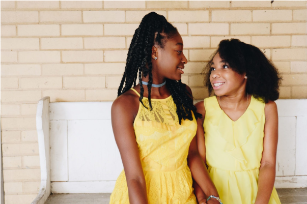 duas mulheres negras usam vestidos amarelos. Elas estão de mãos dadas e sorriem uma para a outra.